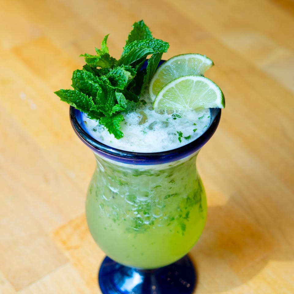 A vibrant green mojito cocktail in a blue glass, garnished with fresh mint and lime slices.