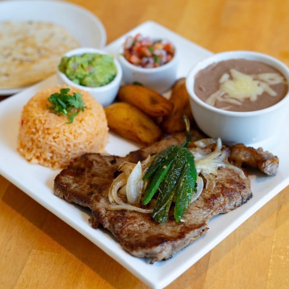 Grilled steak with onions and peppers, rice, beans, guacamole, salsa, and plantains on a white plate.
