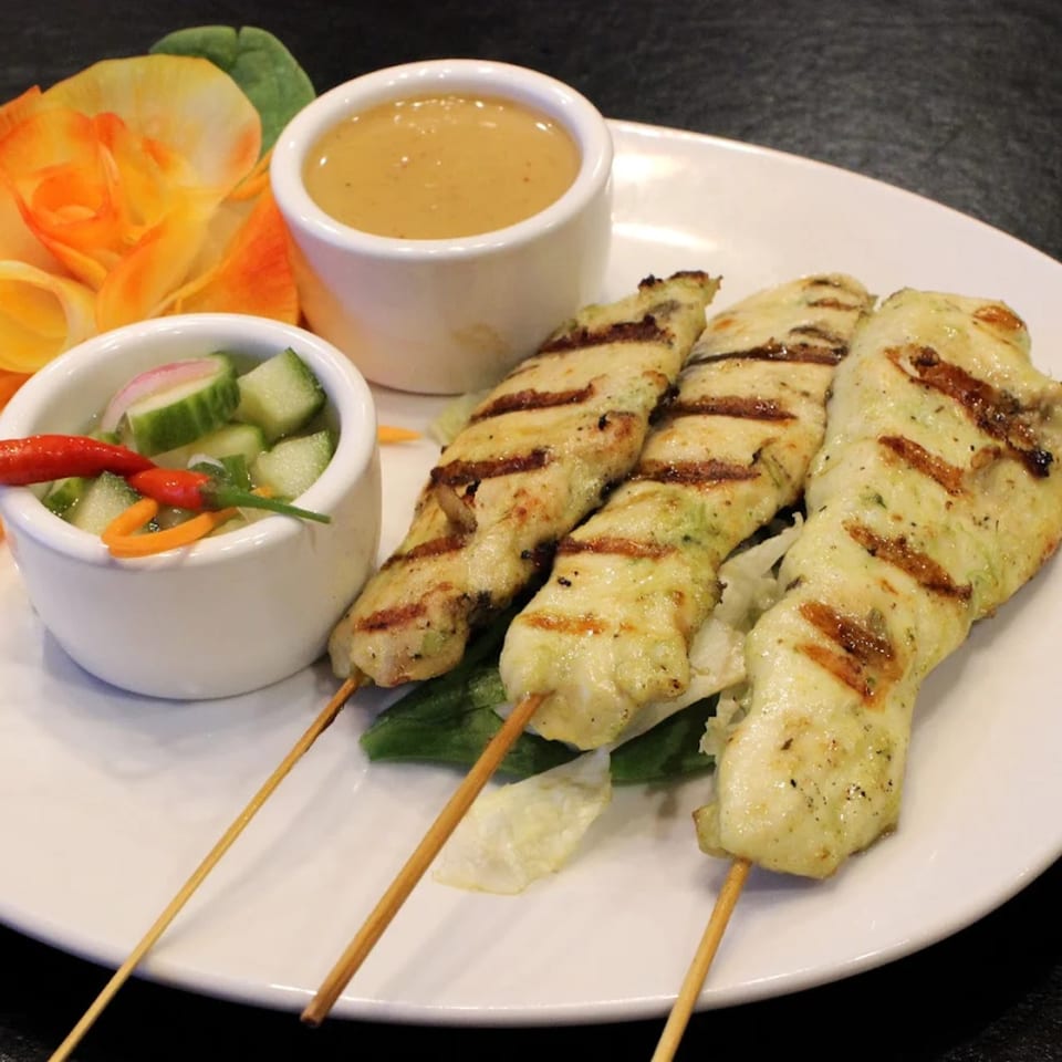 Goji Y Agave's chicken satay skewers with peanut sauce and cucumber salad, garnished with an orange flower.